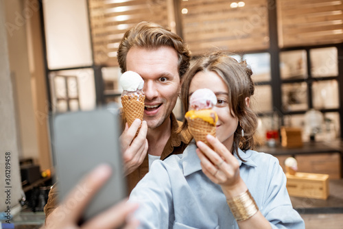 Young and cheerful couple making selfie photo with yummy ice cream in waffle cones, having fun together in the shop or cafe