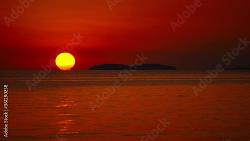 Cinemagraph / seamless video loop of the sun setting over the horizon at a romantic beach with small waves at the Croatian Mediterranean seaside as a big, orange ball with sailing ships passing by.