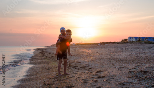 Brother carries a sister on his back along a sandy beach along the sea at sunset. Summer fun. Silhouettes of children on the seashore at sunset. Copy space.
