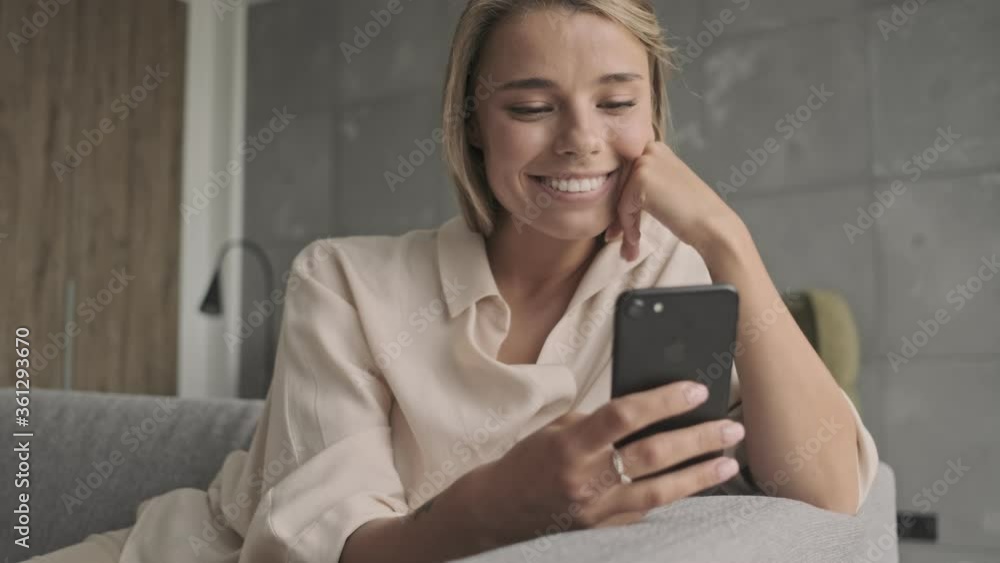 Smiling attractive blonde woman using smartphone and having fun while sitting on sofa at home Smiling attractive blonde woman using smartphone and having fun while sitting on sofa at home