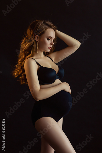 European pregnant woman with long hair in dark wear  happy future mother on black background  young future mom hugs her belly  beautiful wife expecting a baby