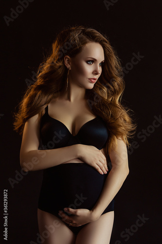 European pregnant woman with long hair in dark wear, happy future mother on black background, young future mom hugs her belly, beautiful wife expecting a baby