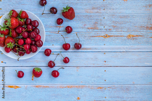 Composition of fresh cherries, strawberries. Bright cherries and strawberries lie on a white plate and a blue wooden background. Summer. Fruits.