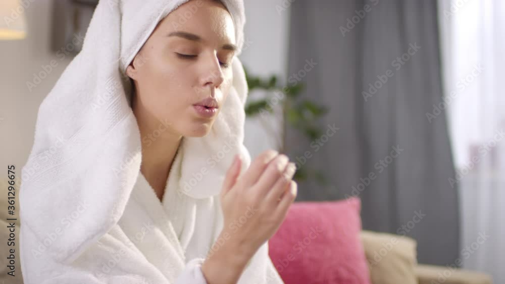 Good-looking young woman wearing white bathrobe applying nail polish and drying it blowing on her fingernails