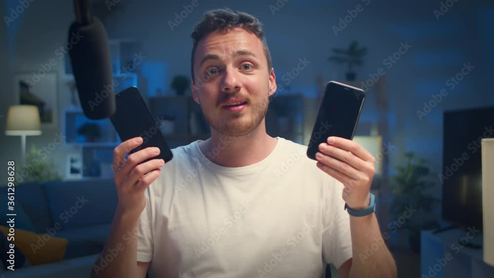 Young handsome blogger reviews two smartphones at home in the living room Young handsome blogger reviews two smartphones at home in the living room