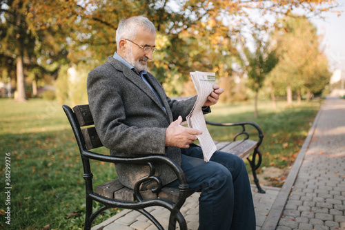Bearded elderly man in glasses reading newspaper in the autumn park. Handsome gray-haired man sitting on the bench early in the morning