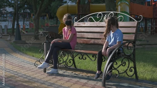 Meet at a distance. Little girls in protective masks sit at opposite ends of the bench and wave their arms to each other from a distance. photo