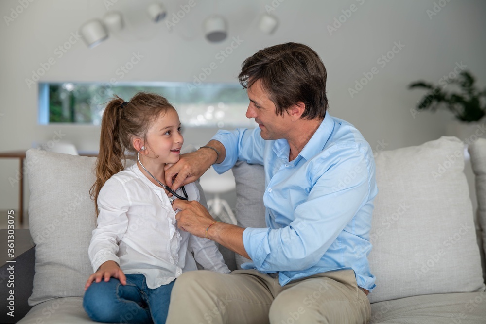 Fototapeta premium Authentic shot of a happy smiling little girl is playing doctor with her father on a sofa in living room at home.