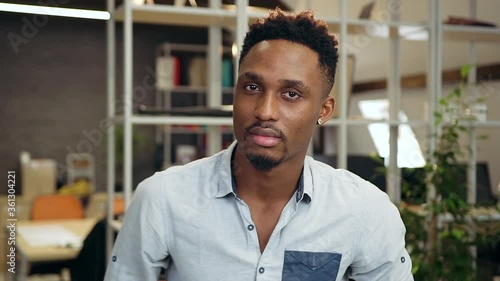 Good-tempered attractive concentrated young dark-skinned guy looking at camera with confident view in the contemporary office photo