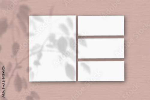 Several horizontal and vertical sheets of white textured paper on the background of a pink wall. Natural light casts shadows from an exotic plant. Flat lay, top view