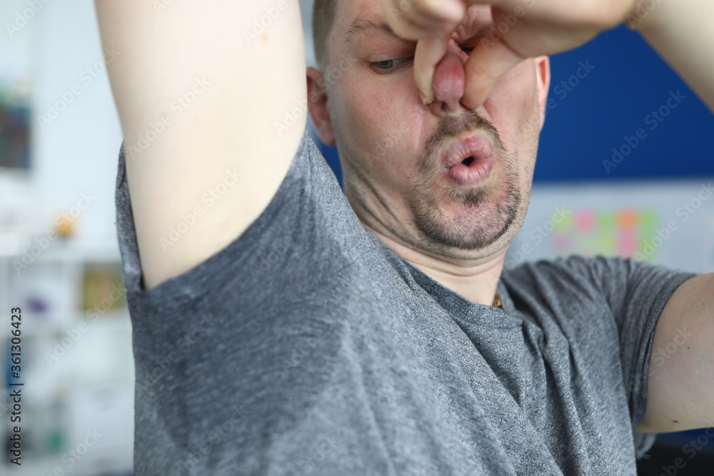 Fototapeta premium Close-up of man after physical exertion. Male person in tee-shirt with sweaty underarm. Sweat on grey shirt. Sportsman plugging nose with hand from stink