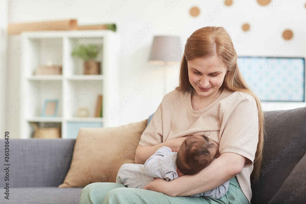 Portrait of happy mature mother breastfeeding baby while sitting on couch in home interior, copy space
