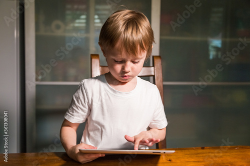 Young girl sitting at the table in the kitchen and watching cartoons on tablet. 