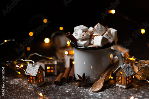 Winter hot drink: hot chocolate with marshmallows, cinnamon and anise. Christmas lights in the shape of houses as decor. Decorative snow, dark wooden background. Festive mood, cozy home atmosphere