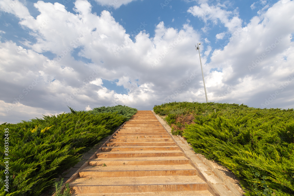 Fototapeta premium Angled closeup of stairs surrounded by greenery, blue sky with white clouds, horizontal