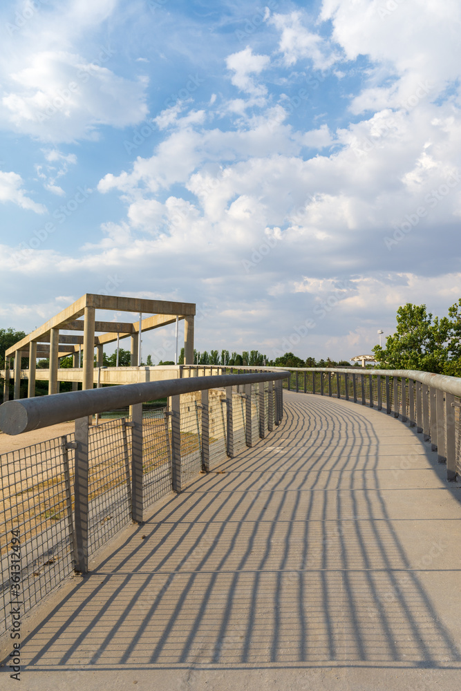 View of path with shadows of fence with concrete bridge in the background, blue sky with white clouds, at sunset, vertical