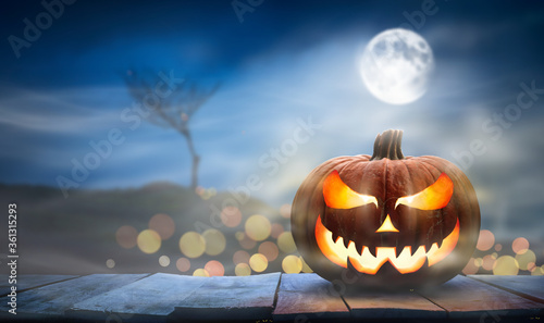 One spooky halloween pumpkin, Jack O Lantern, with an evil face and eyes on a wooden bench, table with a misty night background with space for product placement. photo