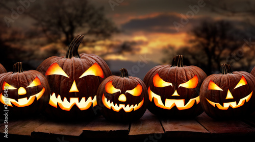 Εκτύπωση καμβά Five spooky halloween pumpkin, Jack O Lantern, with an evil face and eyes on a wooden bench, table with a sunset, night background