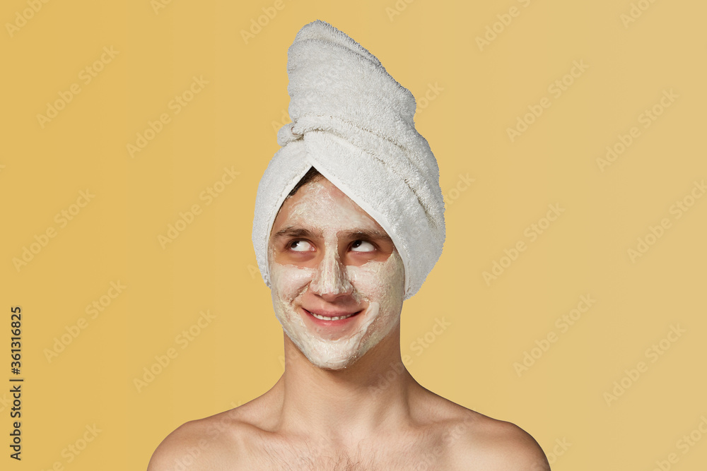 Busy men prefer minimalist, easy and quick facial treatments. A young man with creamy paste-like mask on his face and a white towel on the head is smiling with cunning and looking up. Busy men prefer minimalist, easy and quick facial treatments. A young man with creamy paste-like mask on his face and a white towel on the head is smiling with cunning and looking up.