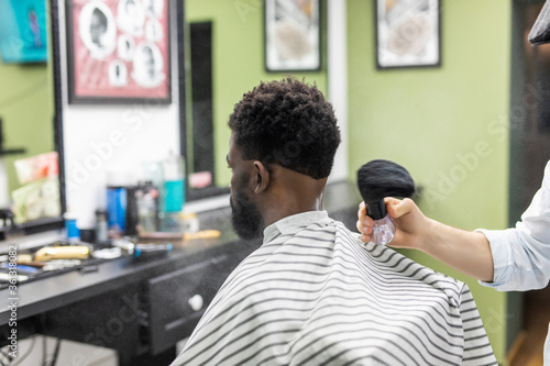 Young black man client getting new haircut in barbershop. Barber hairdresser spreads powder on clients neck with professional shaving brush 