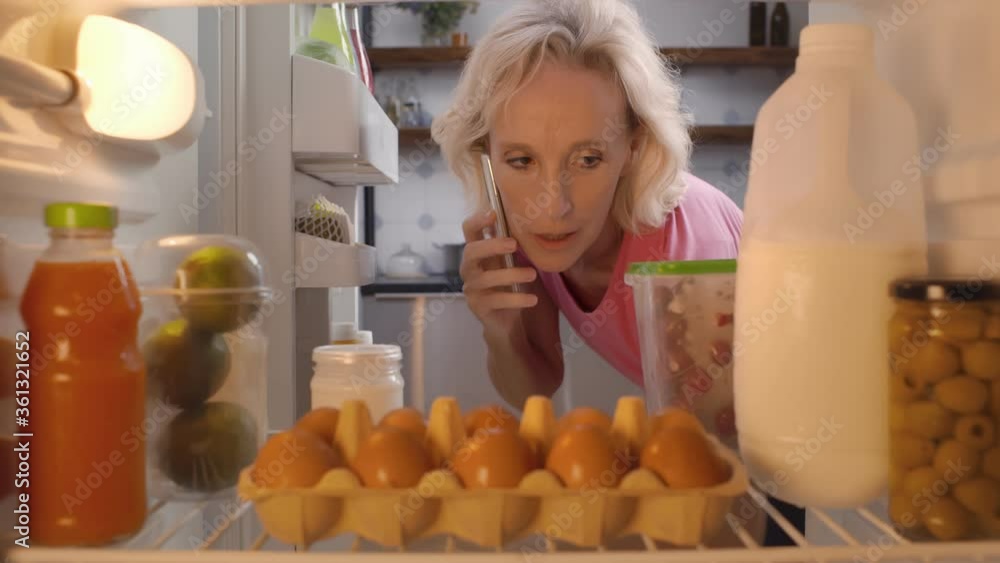 Senior woman talking on phone and taking container with food in fridge Senior woman talking on phone and taking container with food in fridge
