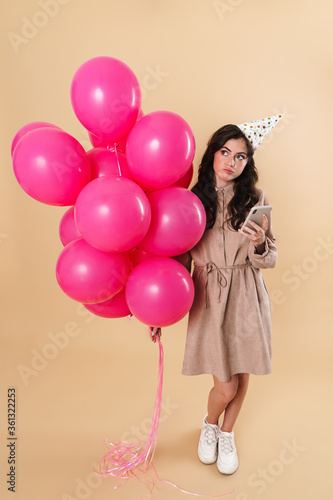 Image of thinking woman using cellphone while posing with pink balloons