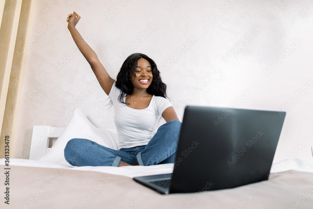 Portrait of smiling young African woman using laptop in bed at home, having video call with her friend, realxing with arm raised up. Good mood on quarantine, lifestyle concept Portrait of smiling young African woman using laptop in bed at home, having video call with her friend, realxing with arm raised up. Good mood on quarantine, lifestyle concept