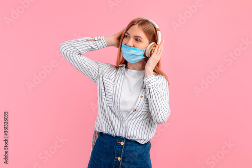 Portrait of a cheerful stylish young woman in a medical protective mask, standing isolated on a pink background, listening to music with headphones