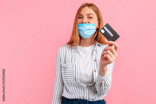 smiling young woman in a medical mask on her face, holding a credit card while rejoicing and looking at the camera on a pink background