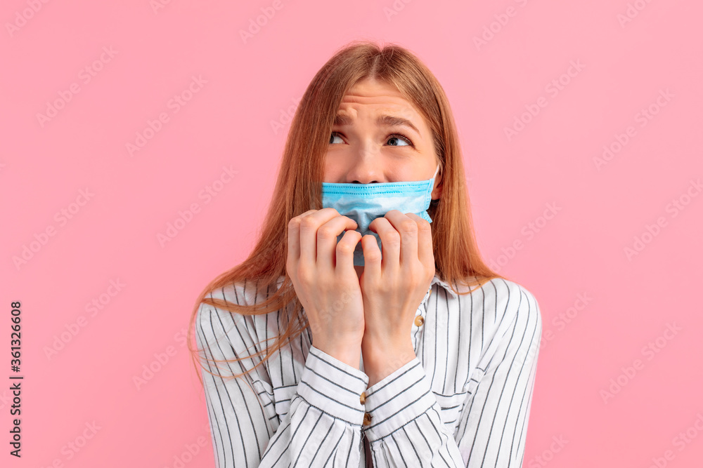 nervous, unsure young woman with a medical mask on her face, looking away, feeling fear, biting her nails with fear, on a pink background nervous, unsure young woman with a medical mask on her face, looking away, feeling fear, biting her nails with fear, on a pink background