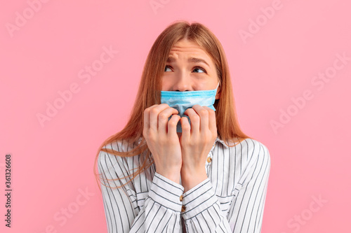 nervous, unsure young woman with a medical mask on her face, looking away, feeling fear, biting her nails with fear, on a pink background
