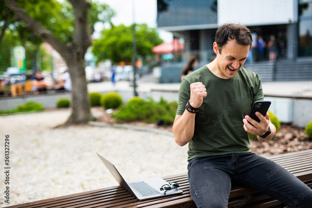 Obraz premium Happy young man celebrate win look at phone sitting on a bench in the park