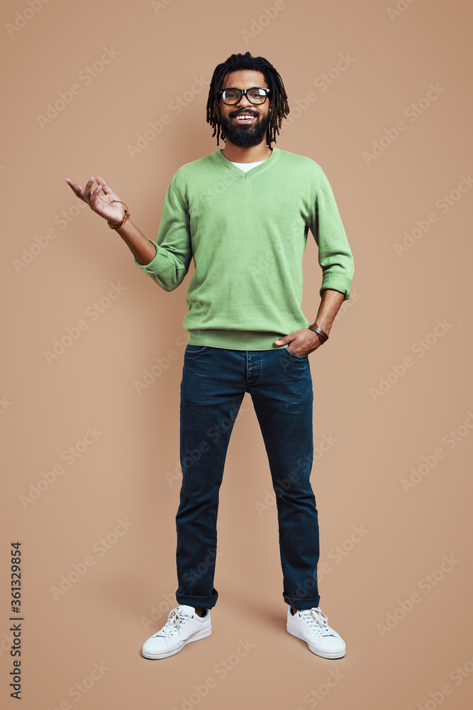 Full length of young African man in casual clothing pointing copy space and smiling while standing against brown background Full length of young African man in casual clothing pointing copy space and smiling while standing against brown background