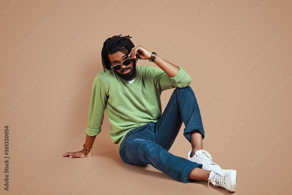 Trendy young African man in casual clothing looking at camera and adjusting eyewear while sitting against brown background Trendy young African man in casual clothing looking at camera and adjusting eyewear while sitting against brown background