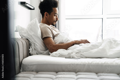 Photo of african american man working with laptop while lying in bed