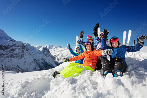 Group of kids in colorful ski sport snow outfit rise hands sitting together over mountain tops
