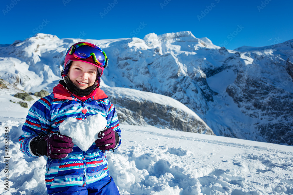Ski little girl hold snow in heart shape smiling with mountain summit on background