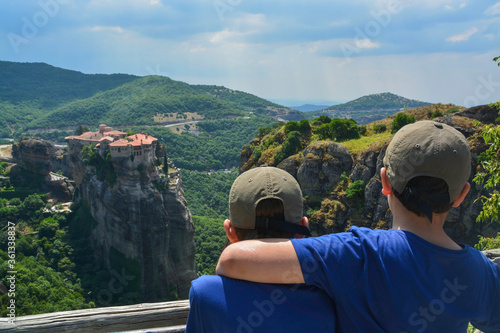 
Varlaam Monastery, Meteora, Greece, Summer 2019. Two boys with their backs to each other in caps at the viewpoint of the Grand Meteora monastery watching the Varlaam monastery
