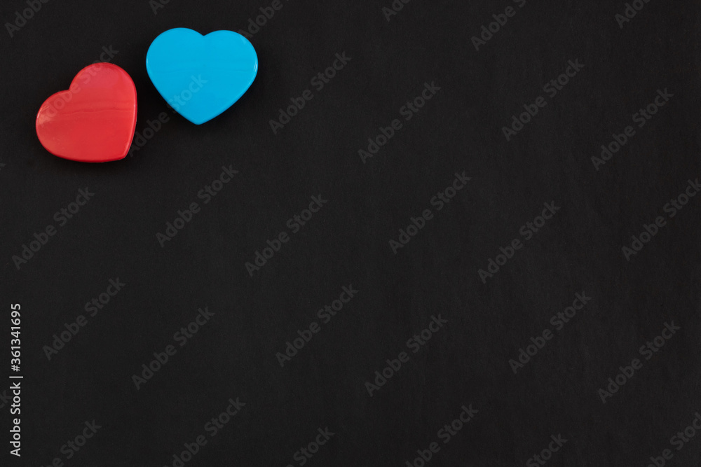 Two red and blue color hearts lying close together isolated at the top left corner of black paper background. Flat lay with empty space for text. Two red and blue color hearts lying close together isolated at the top left corner of black paper background. Flat lay with empty space for text.
