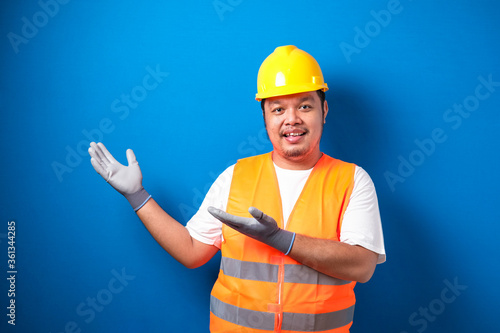 Fat Asian worker wearing a helmet presents something in his hand while facing sideways. the man showed an expression of joy and welcome against blue background