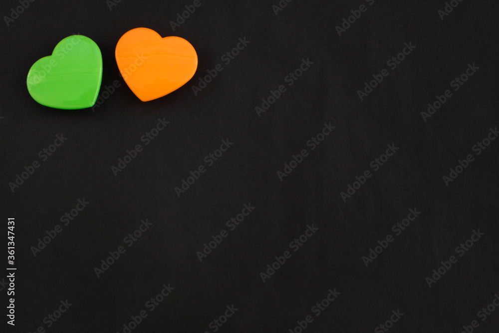 Fototapeta premium Two green and orange color hearts lying close together isolated at the top left corner of black paper background. Flat lay with empty space for text.
