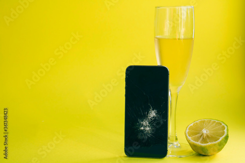 Broken black smartphone with a glass of champagne and lemon on a bright yellow background