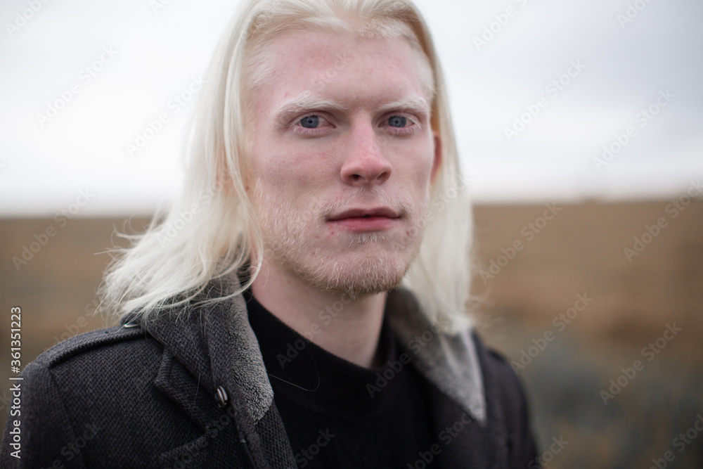 Portrait of a young, beautiful albino man with blue eyes and shoulder hair in a black quilted hooded coat, looking at the camera with a shrewd look against the background of the field. Portrait of a young, beautiful albino man with blue eyes and shoulder hair in a black quilted hooded coat, looking at the camera with a shrewd look against the background of the field.