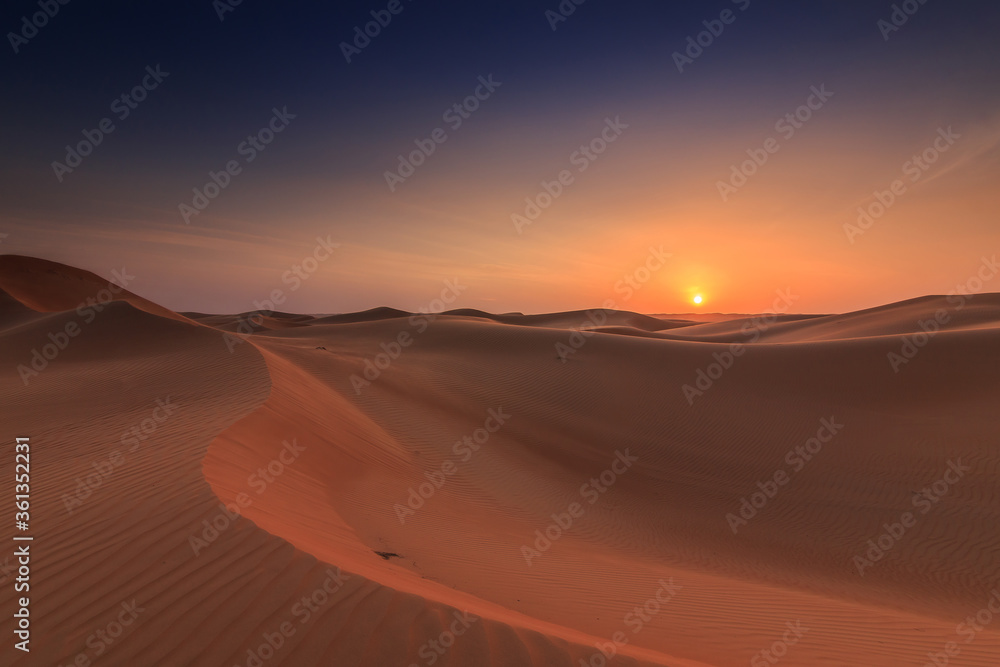 Sunset in the wahiba sands desert of the Sultanate of Oman. Untouched and endless dunes in the evening glow orange from the evening sun. Blue sky with sun and few clouds on the horizon