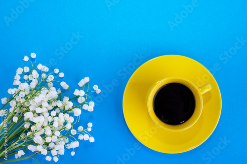 Yellow cup with coffee on a blue background. Lily of the valley flowers