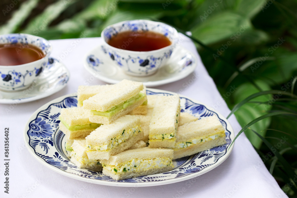 Afternoon tea in the garden, finger sandwiches with cucumber and egg salad and tea on a light background.