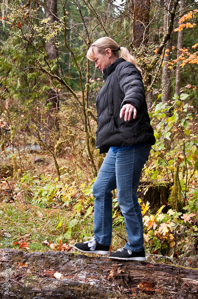 Fototapeta premium This midlife aged Caucasian woman is balancing while walking on a log in the forest on a cold, damp day. She's wearing blue jeans, and a black down coat.