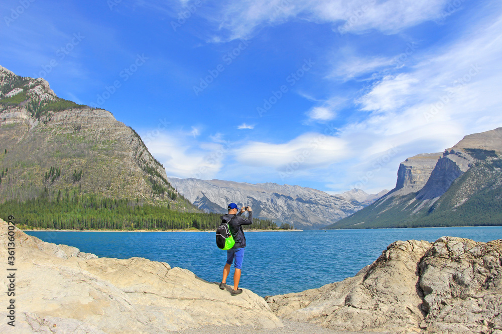 Backpacker / man / male hiker standing on the edge of rocky cliff, taking picture of Minnewanka lake in Rocky mountains -  Banff National Park, AB