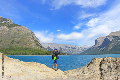 Backpacker / man / male hiker standing on the edge of rocky cliff, taking picture of Minnewanka lake in Rocky mountains - Banff National Park, AB