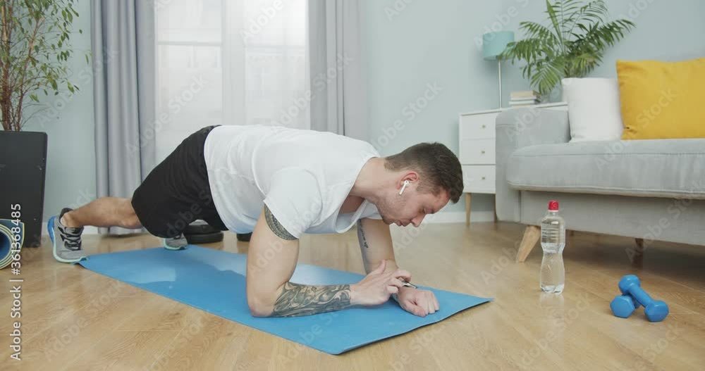 A muscular trainer practicing sports and working out. A young man doing plank exercise using a smartwatch device. In-home training during pandemic, quarantine, lockdown, isolation period. Stay home. A muscular trainer practicing sports and working out. A young man doing plank exercise using a smartwatch device. In-home training during pandemic, quarantine, lockdown, isolation period. Stay home.